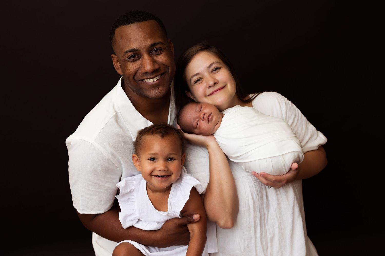 A loving family portrait against a black background shows a group in white attire embracing and smiling together.