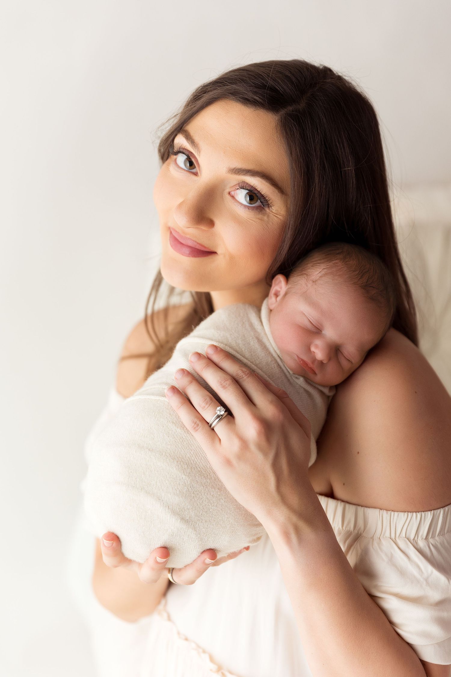 Mother in white top holds sleeping swaddled newborn against white background.