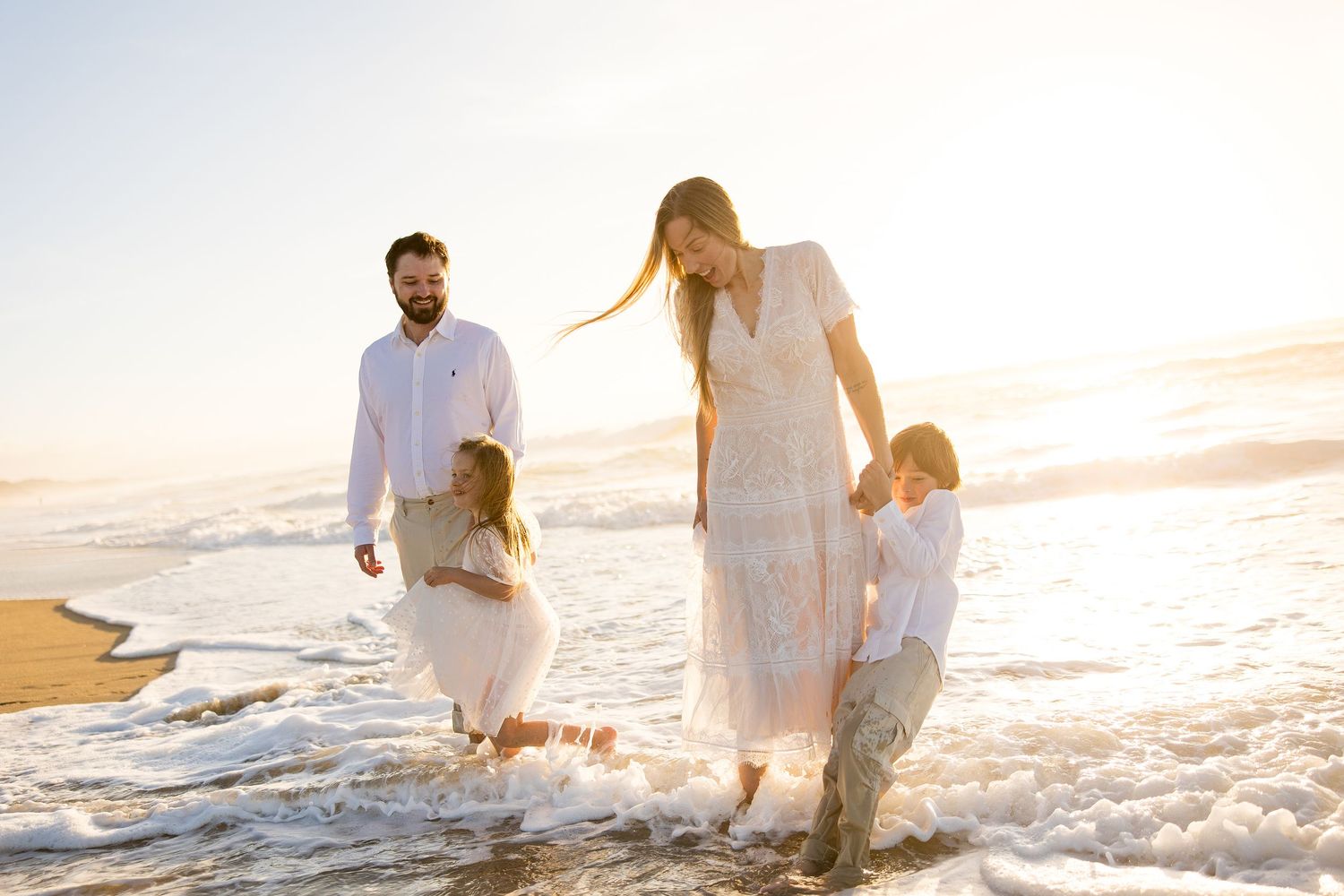 A couple in white outfits walks along a sandy Marina California beach at sunset while holding hands.