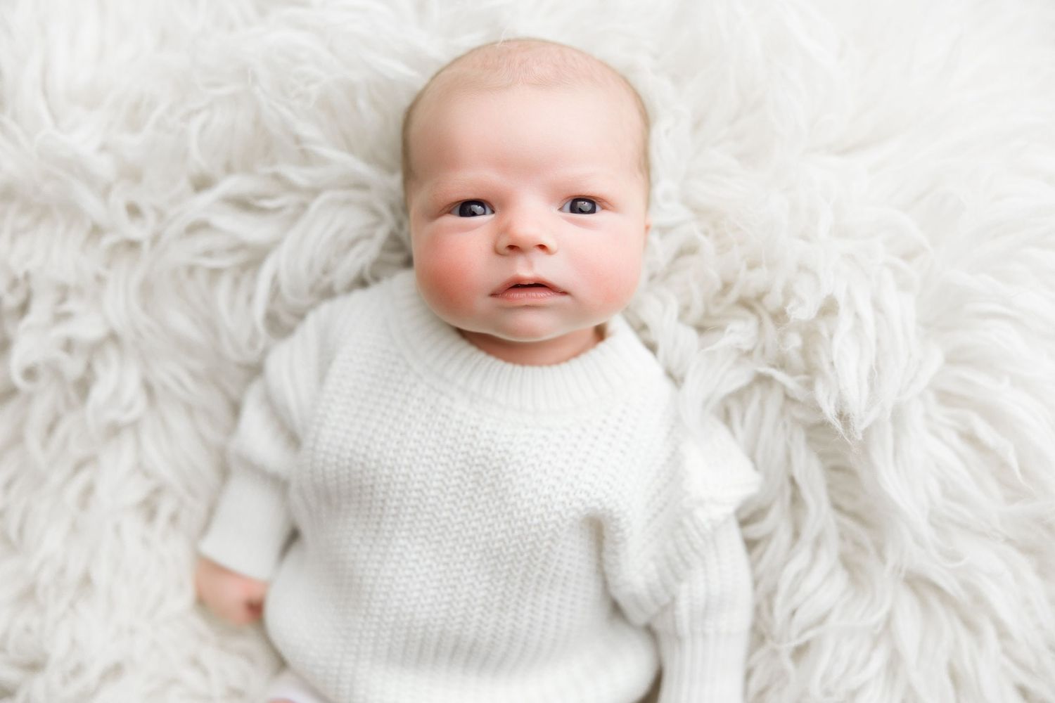 A baby wearing a white knit sweater lies on a soft white fur blanket while looking at the camera.