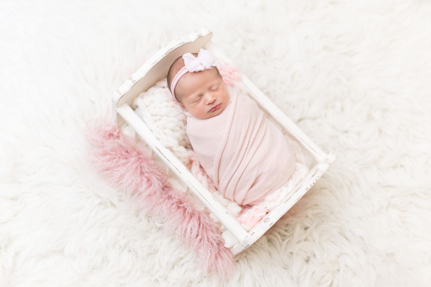 A newborn baby sleeps peacefully on soft white fur while surrounded by pink decorative accents.