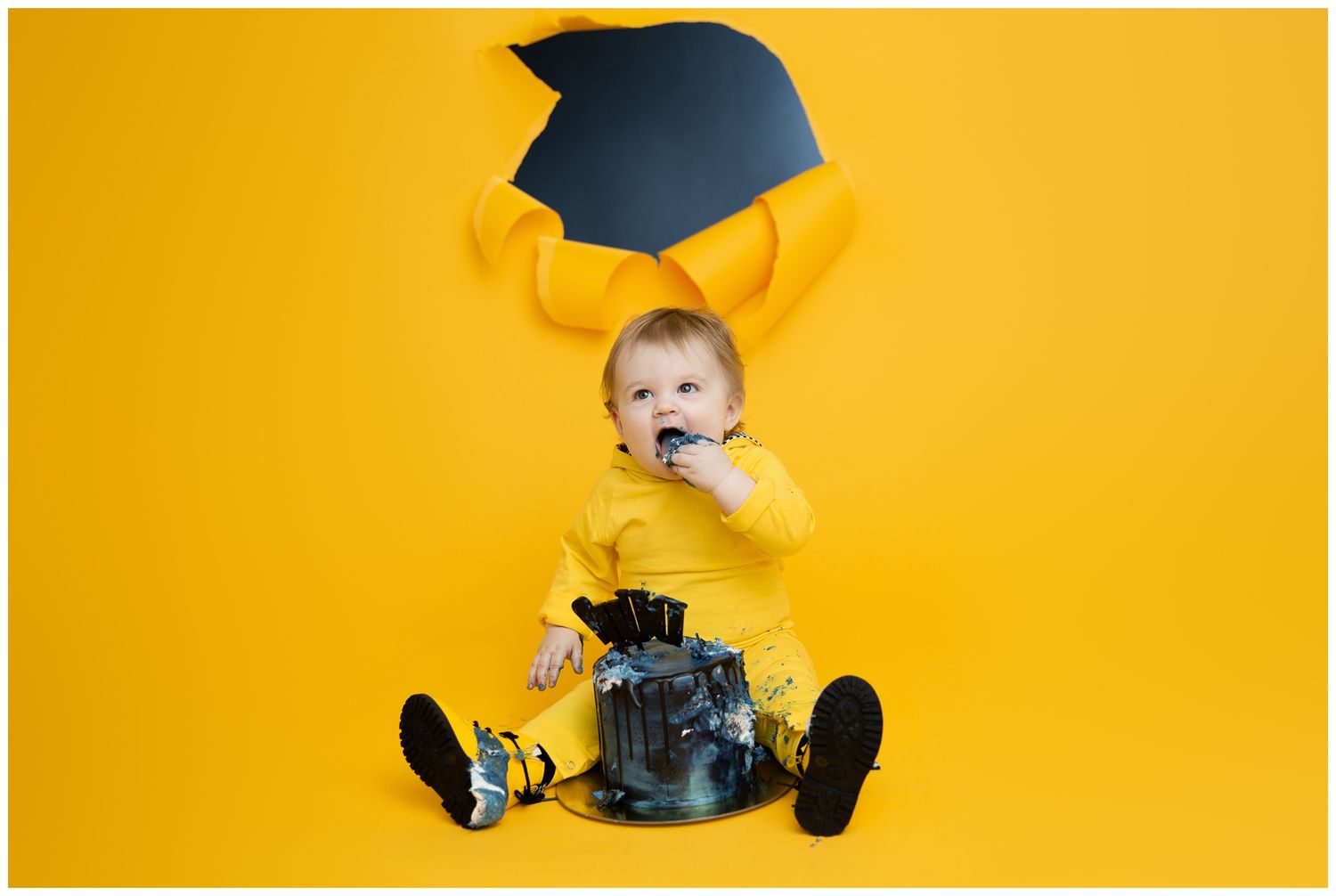 Baby in a black graduation cap and yellow shoes sits against a bright yellow background.