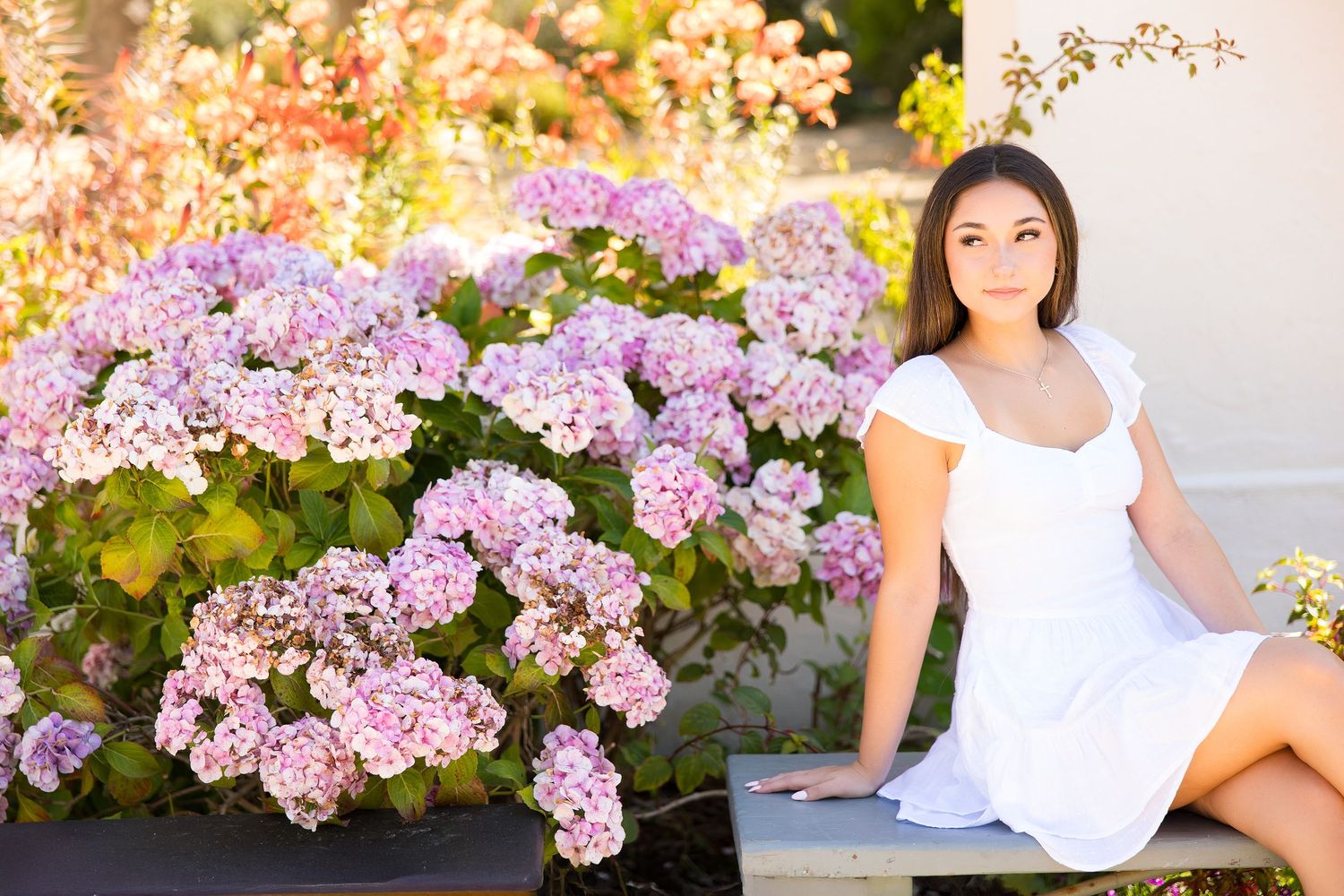 Young woman in white dress sitting beside blooming pink and purple hydrangeas in a sunlit garden.