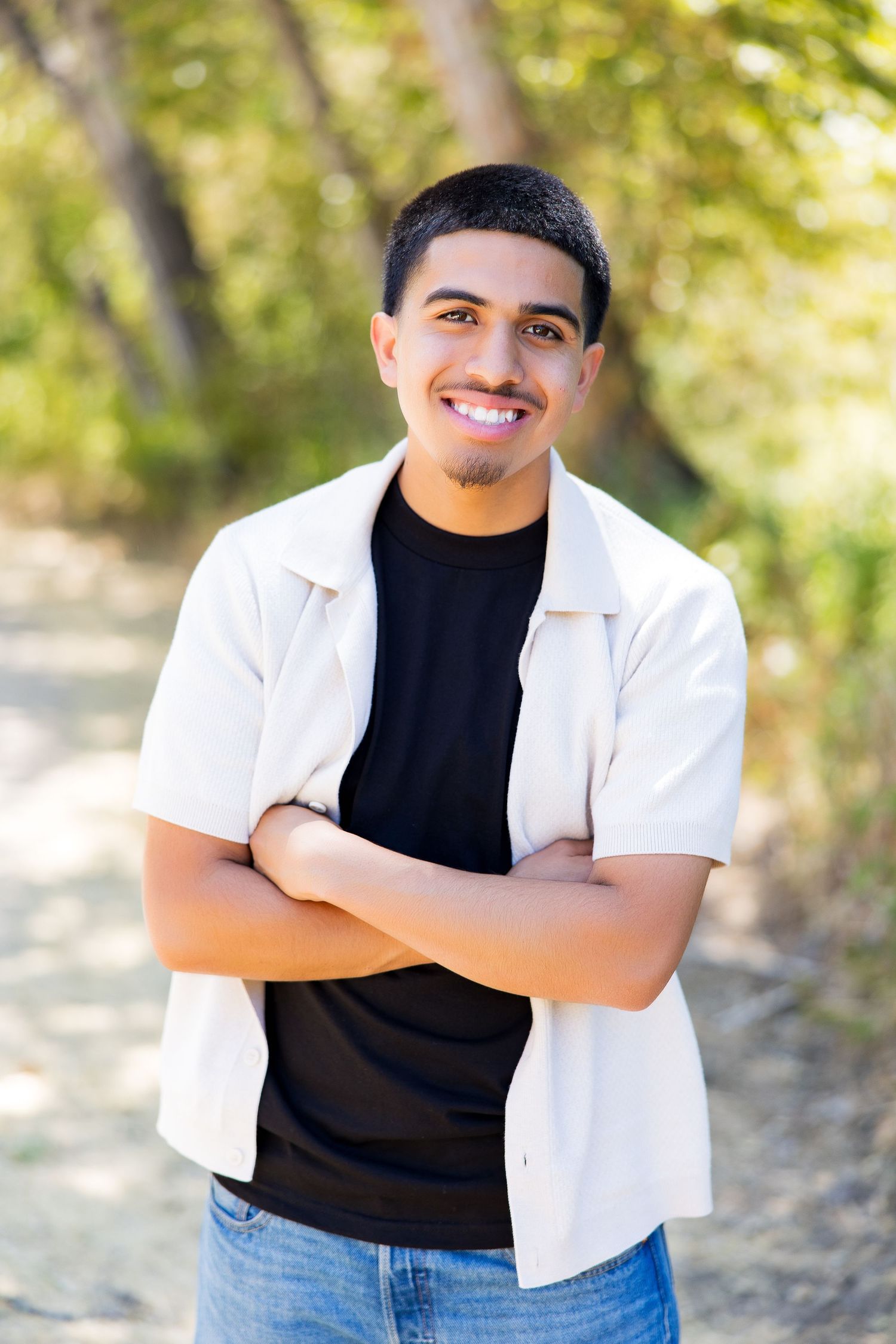 Smiling young Hispanic man standing outdoors with arms crossed, wearing white button shirt over black tee and jeans.