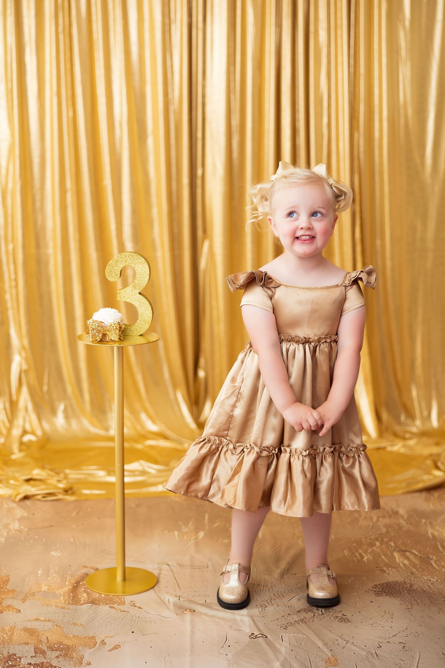 Toddler wearing a beige dress standing against a shimmering gold curtain backdrop.