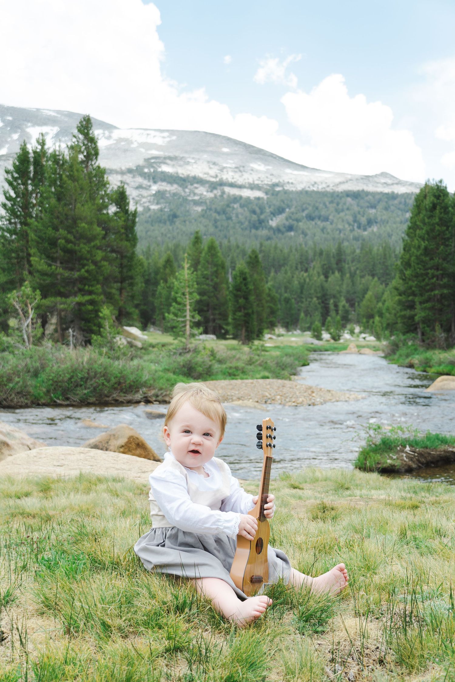 A small child sits with a guitar by a mountain stream surrounded by evergreen trees, sound of music inspired in Yosemite.
