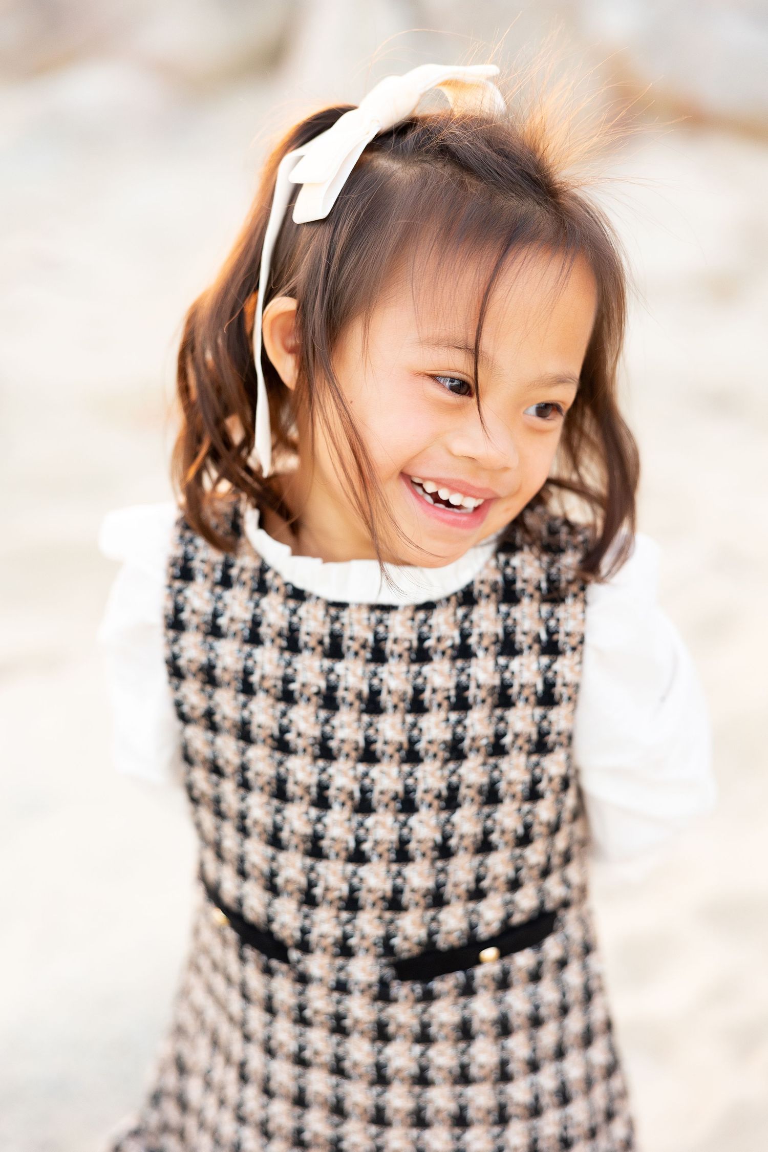 Close up portrait of child in houndstooth dress and white bow laughing on beach.
