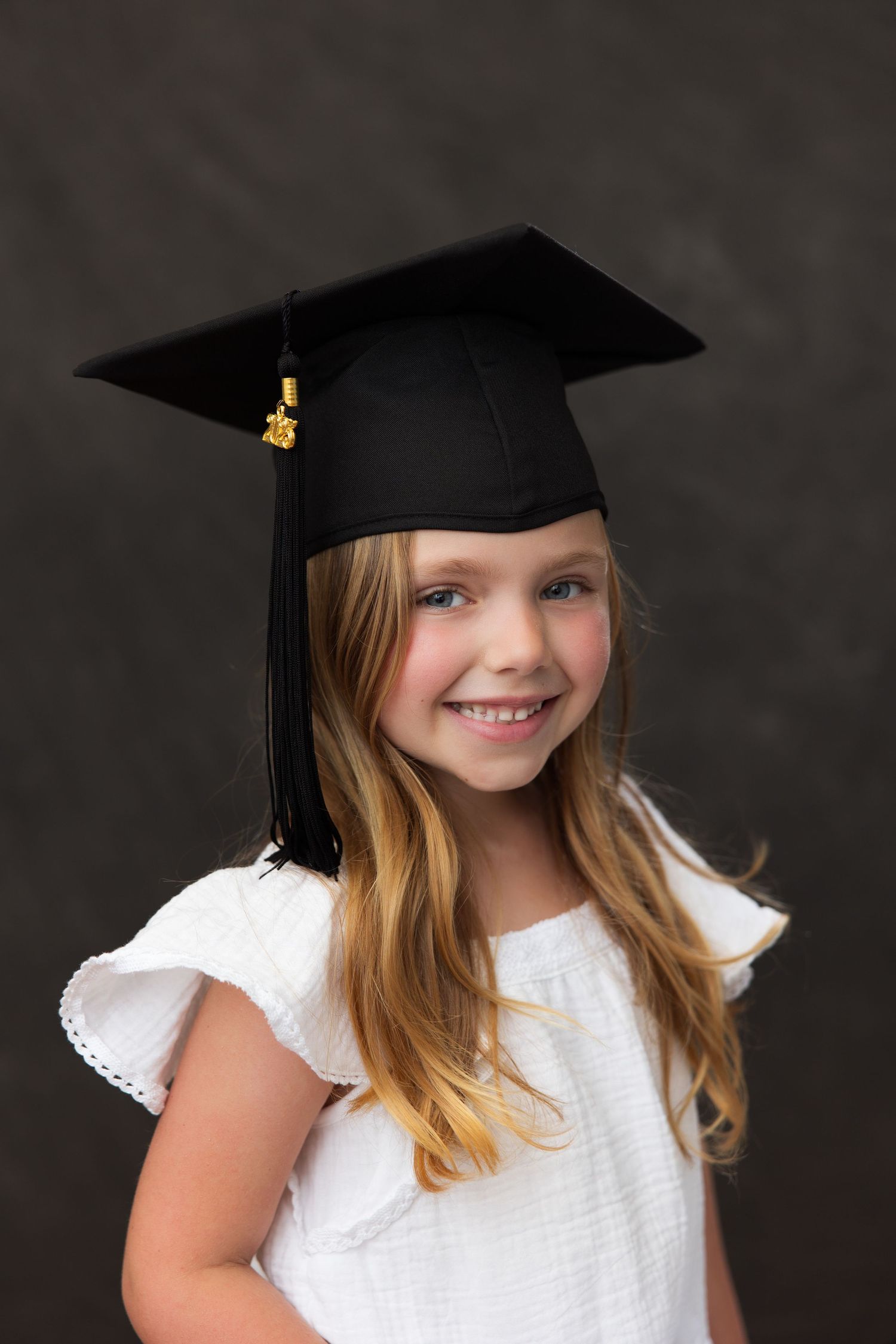 A young graduate wearing a black mortarboard cap and white dress smiles during a portrait session against a dark background.