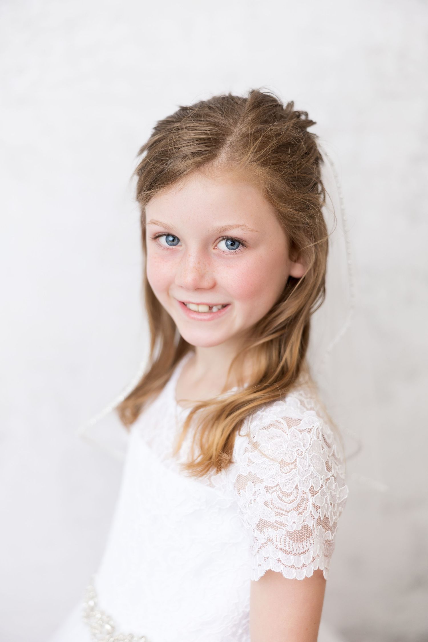 A child in a white lace dress with blonde hair smiles against a light background for a portrait.