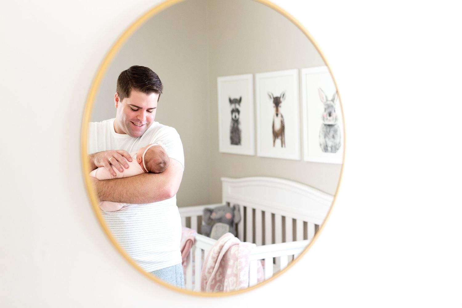 Mirror reflection shows parent holding baby in neutral toned nursery with animal artwork.