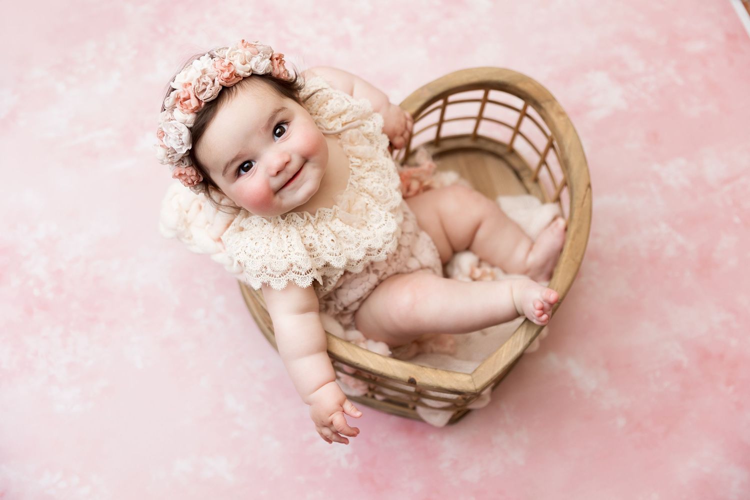 Baby in vintage lace collar rests in wicker basket against pink backdrop.