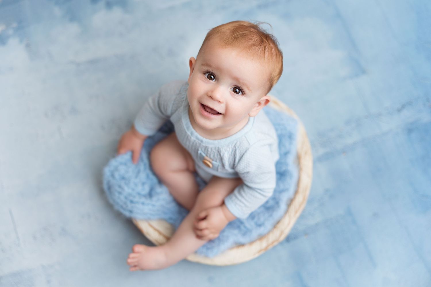 Baby in light blue outfit sitting on round cushion against soft blue backdrop.