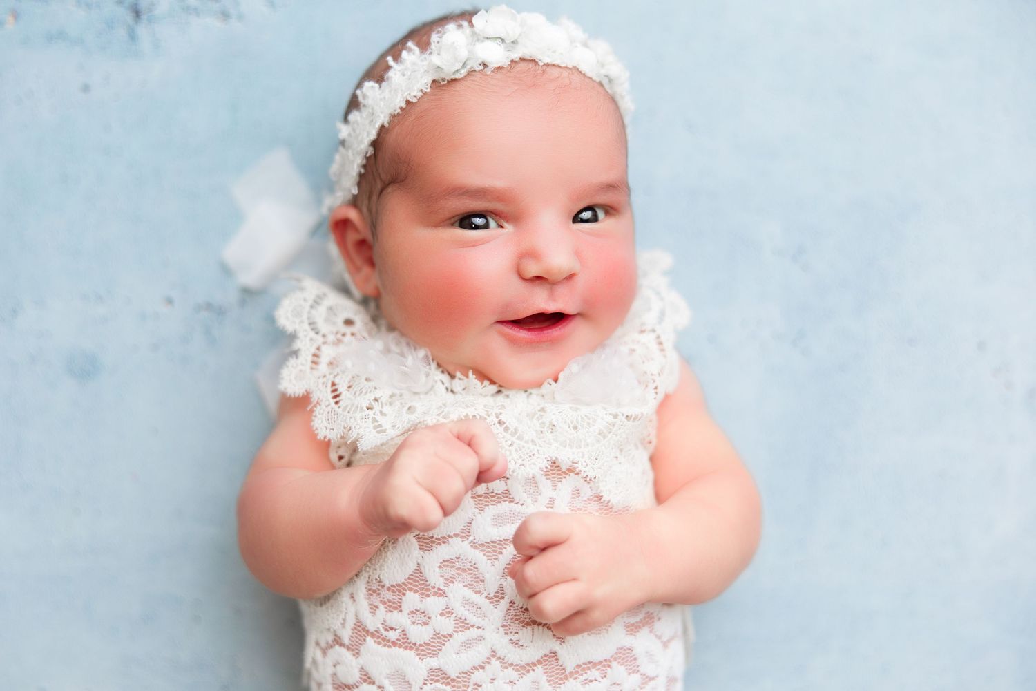 Baby in white lace dress and headband against light blue background.