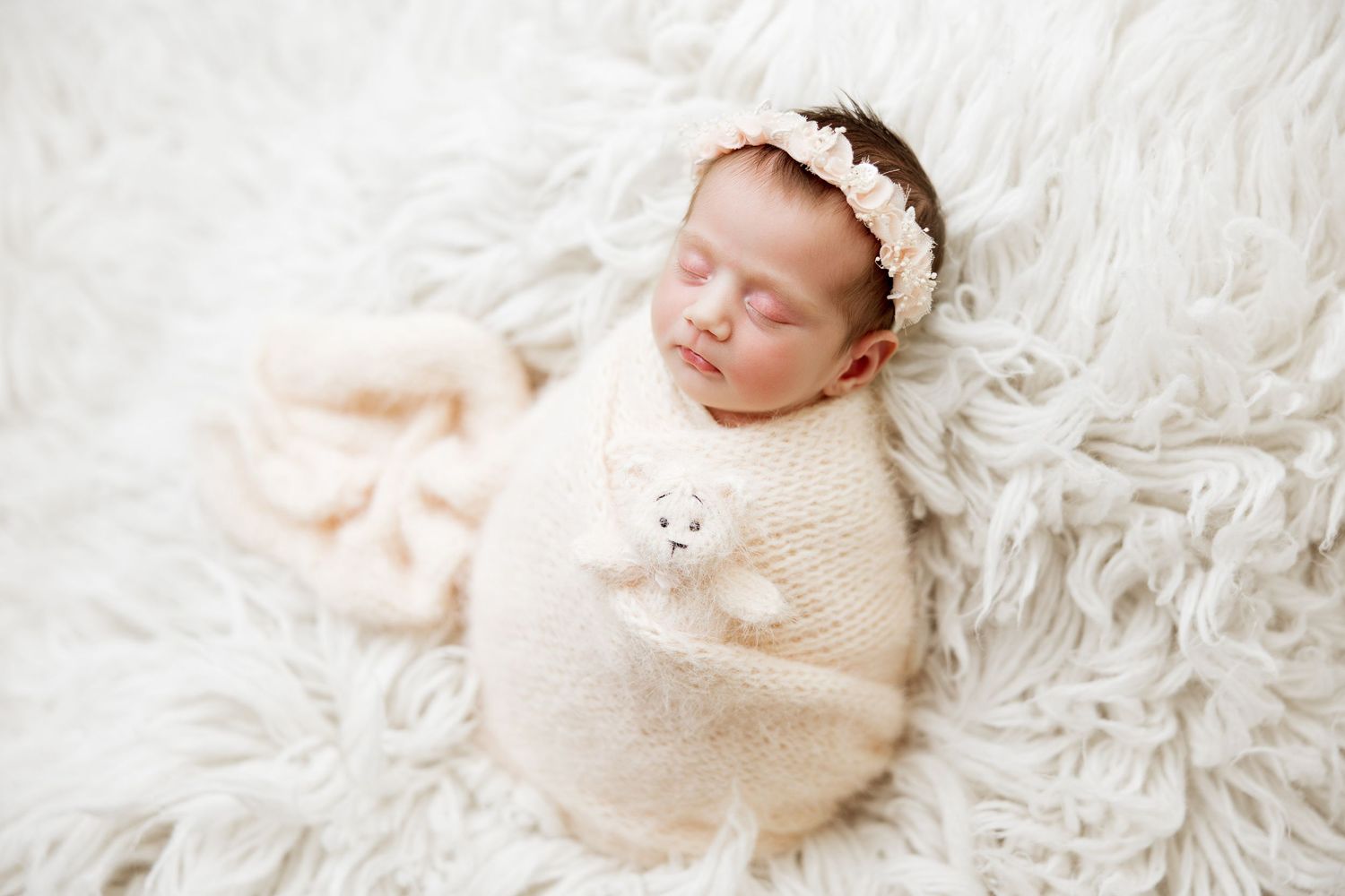 Sleeping newborn wrapped in cream swaddle with headband on white furry backdrop.
