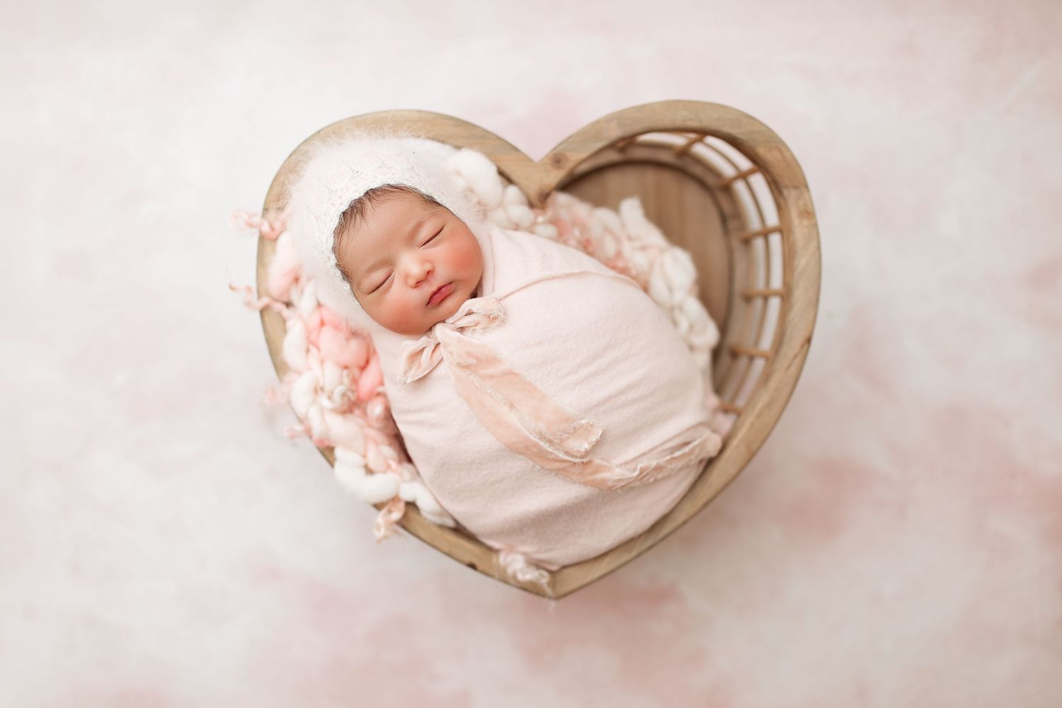 Newborn baby peacefully sleeping in heart-shaped basket on pink background.