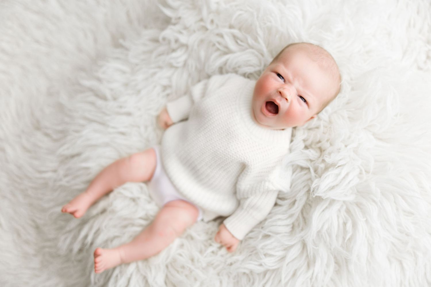 A baby in a white onesie lies on a plush white fur rug while yawning with arms outstretched.
