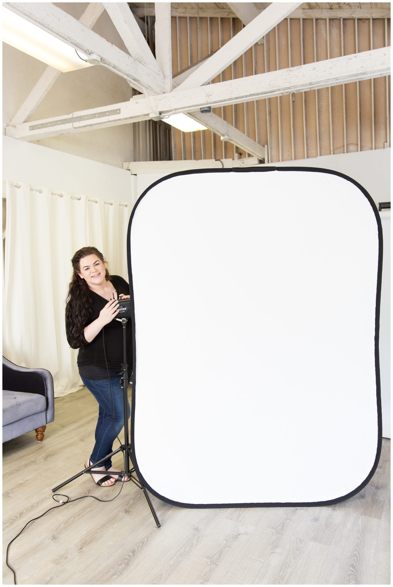 A photography setup in a room with exposed ceiling beams shows a large white reflector screen on a stand next to furniture.