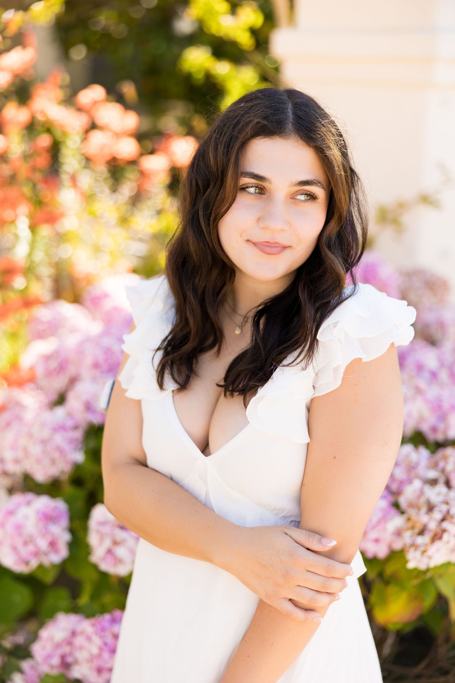 Young woman in white ruffled dress posing with arms crossed among colorful garden flowers.