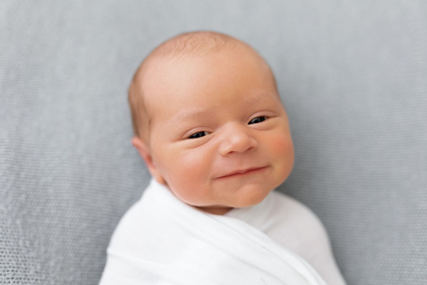 A smiling baby lies on a soft gray blanket showing a progression of happy expressions.