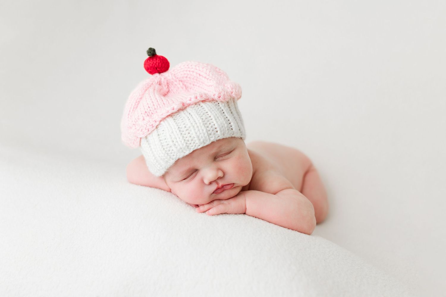 Sleeping newborn baby wearing pink knit hat rests peacefully on white blanket during professional photo session.
