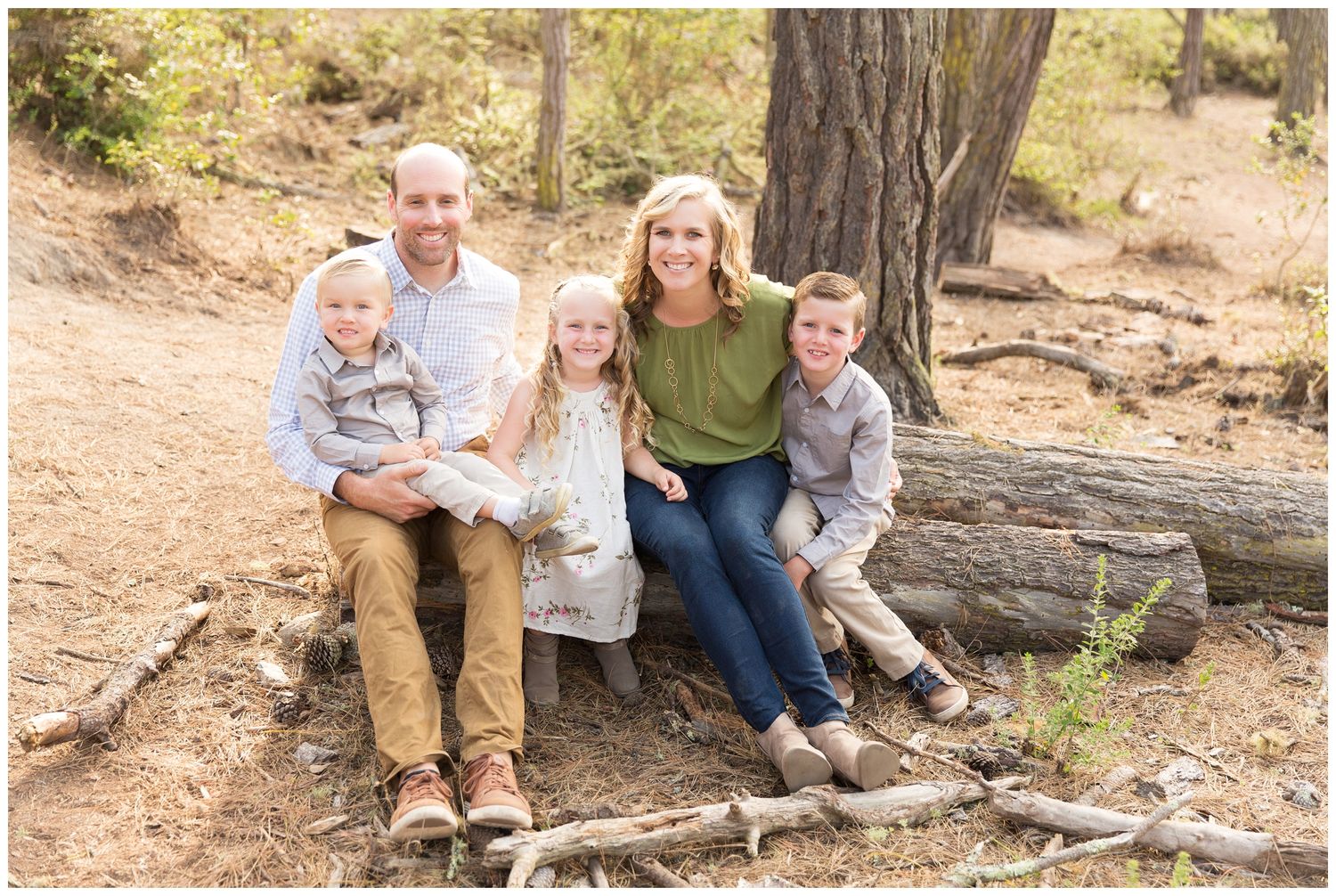 A family sits together on a fallen log in a sunny wooded area during an outdoor photo session.