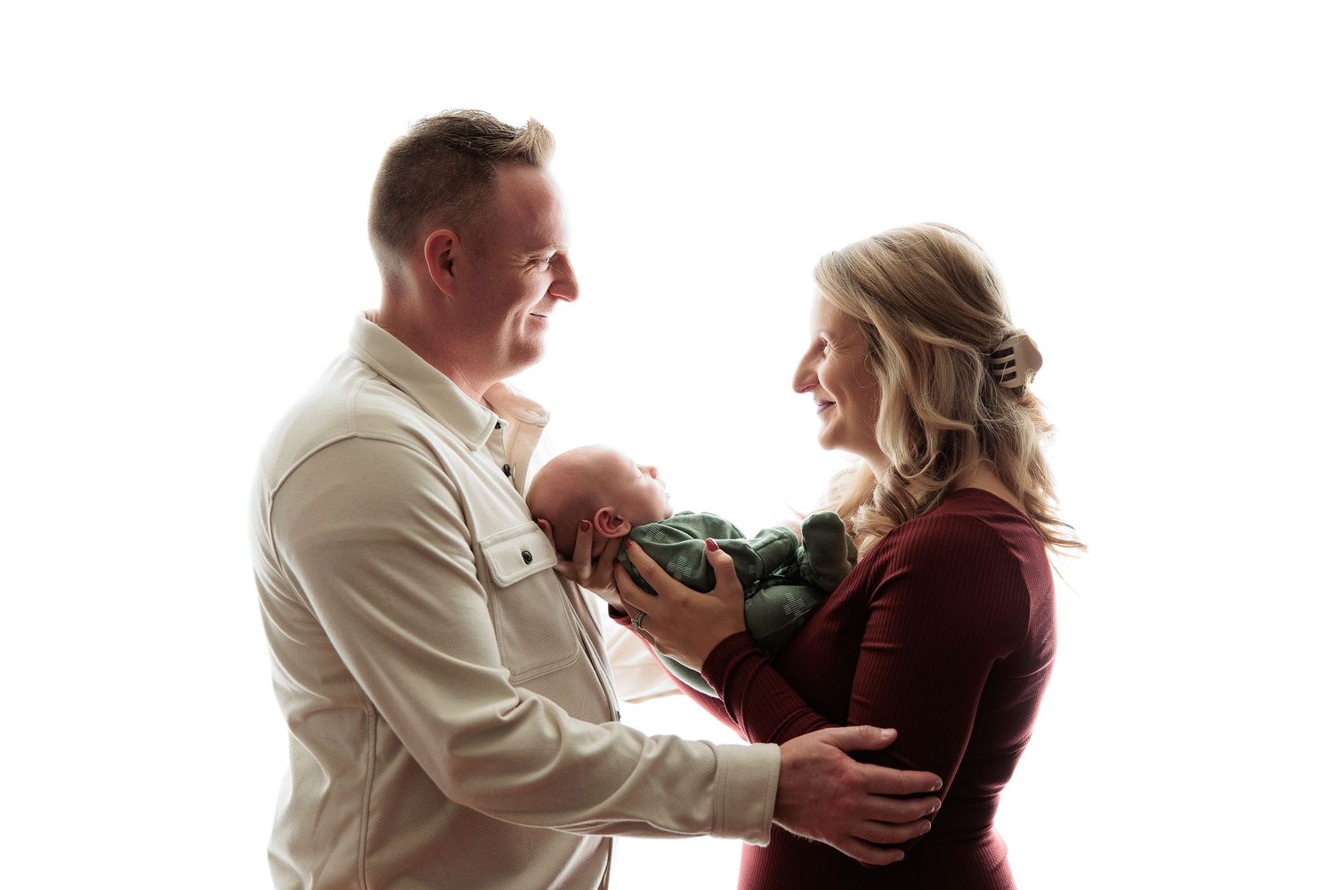 A couple in white and burgundy holds their newborn baby against a bright white studio backdrop.