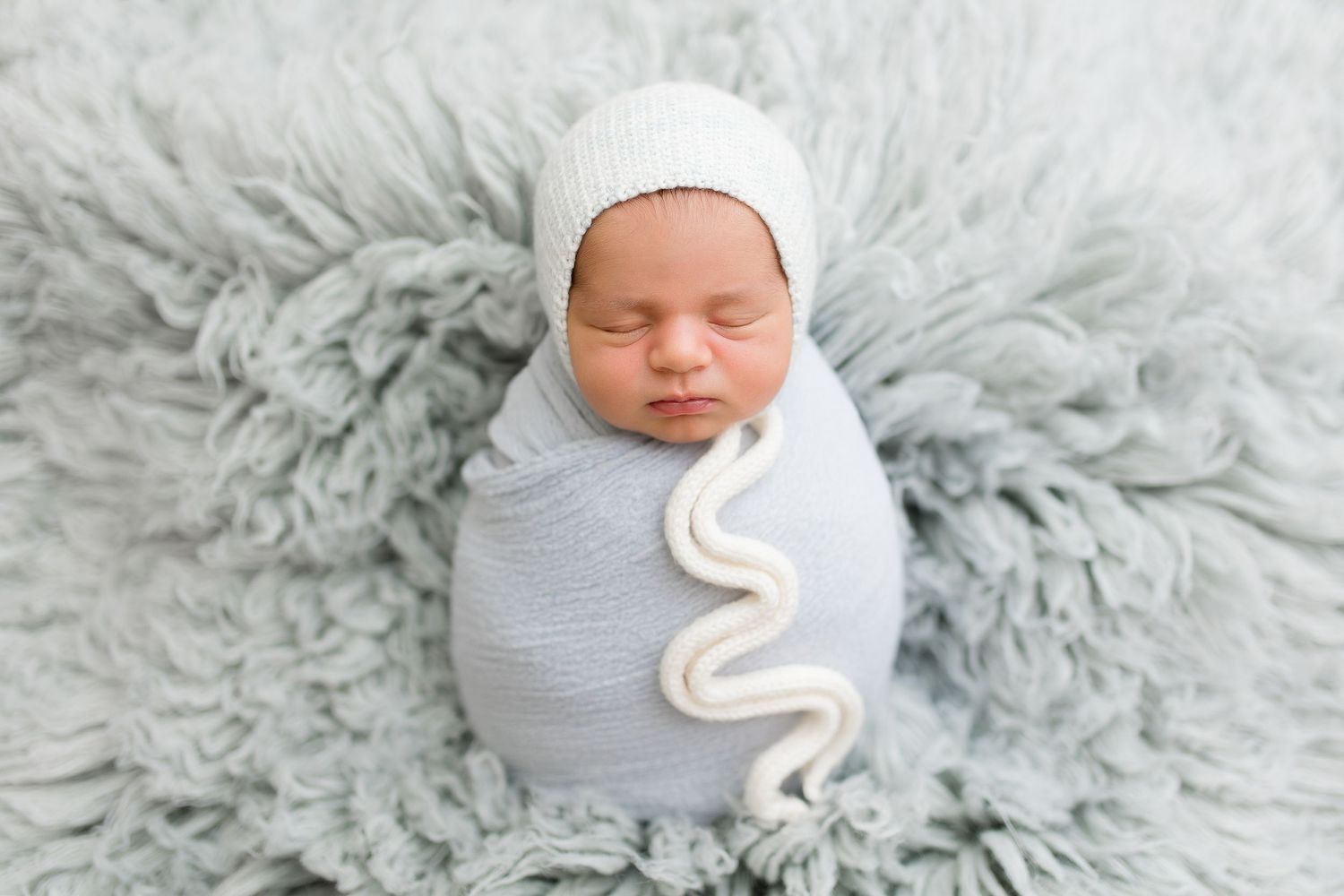 Sleeping newborn swaddled in white wrap lies peacefully on fluffy gray blanket.