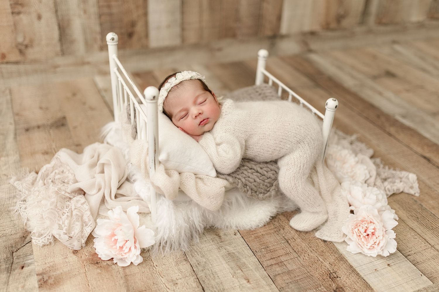 Peaceful sleeping newborn in white outfit laying on miniature bed with roses on wooden floor.