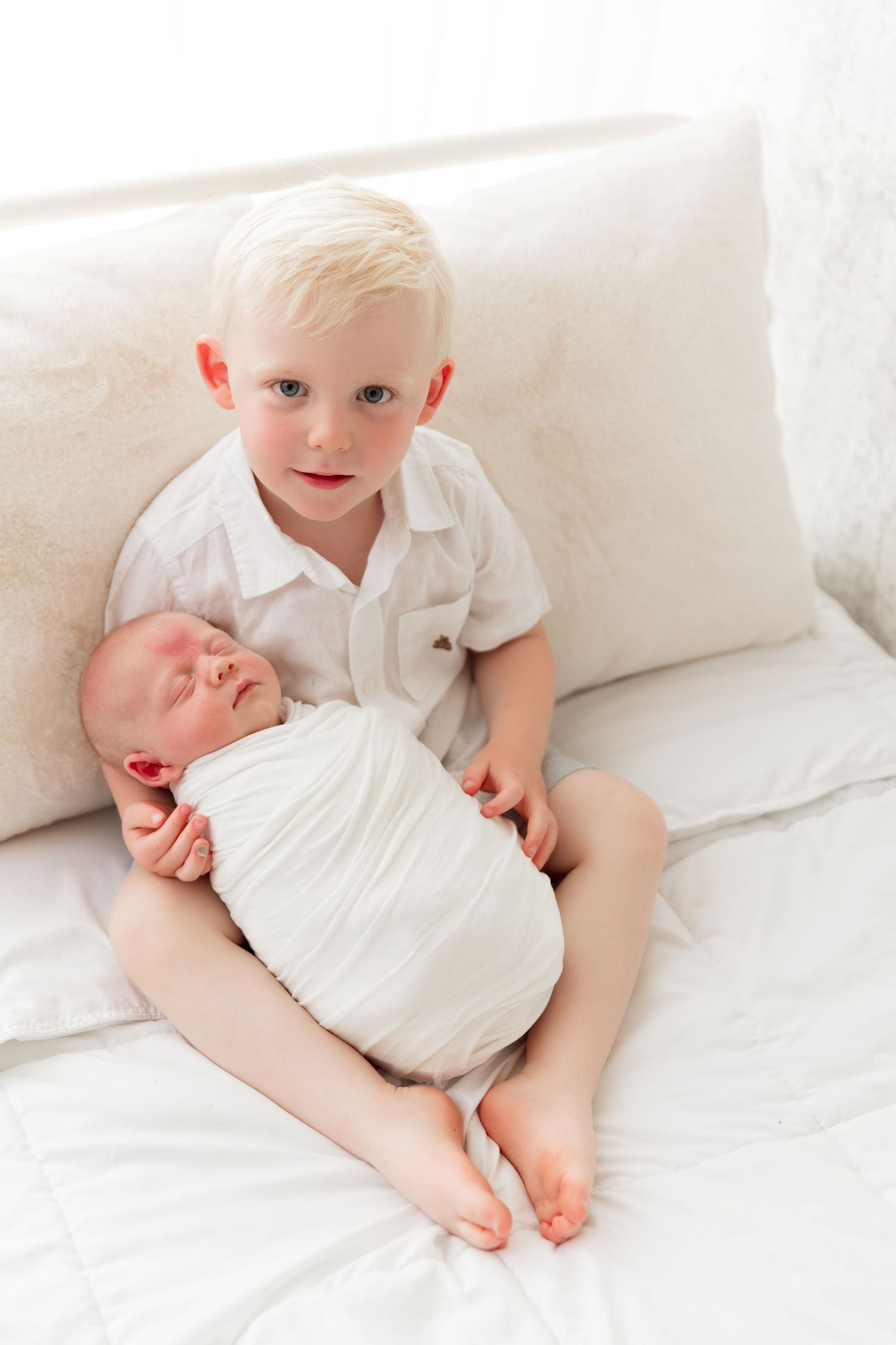 Siblings sitting together on white couch with newborn.