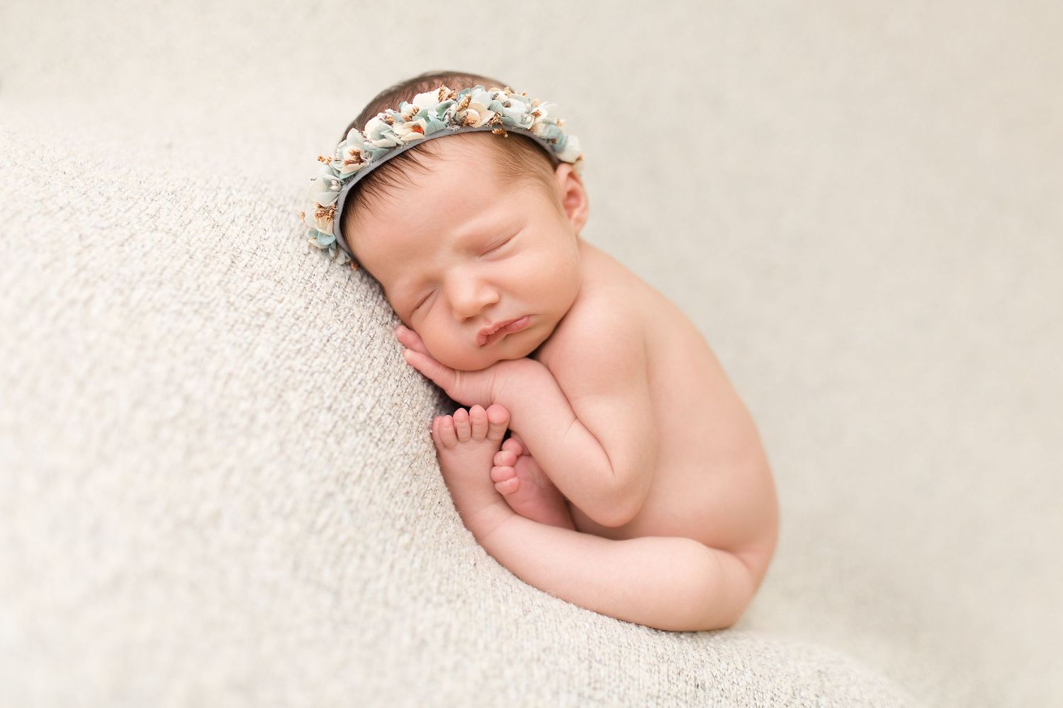 Peaceful sleeping newborn wearing floral crown on cream textured backdrop.