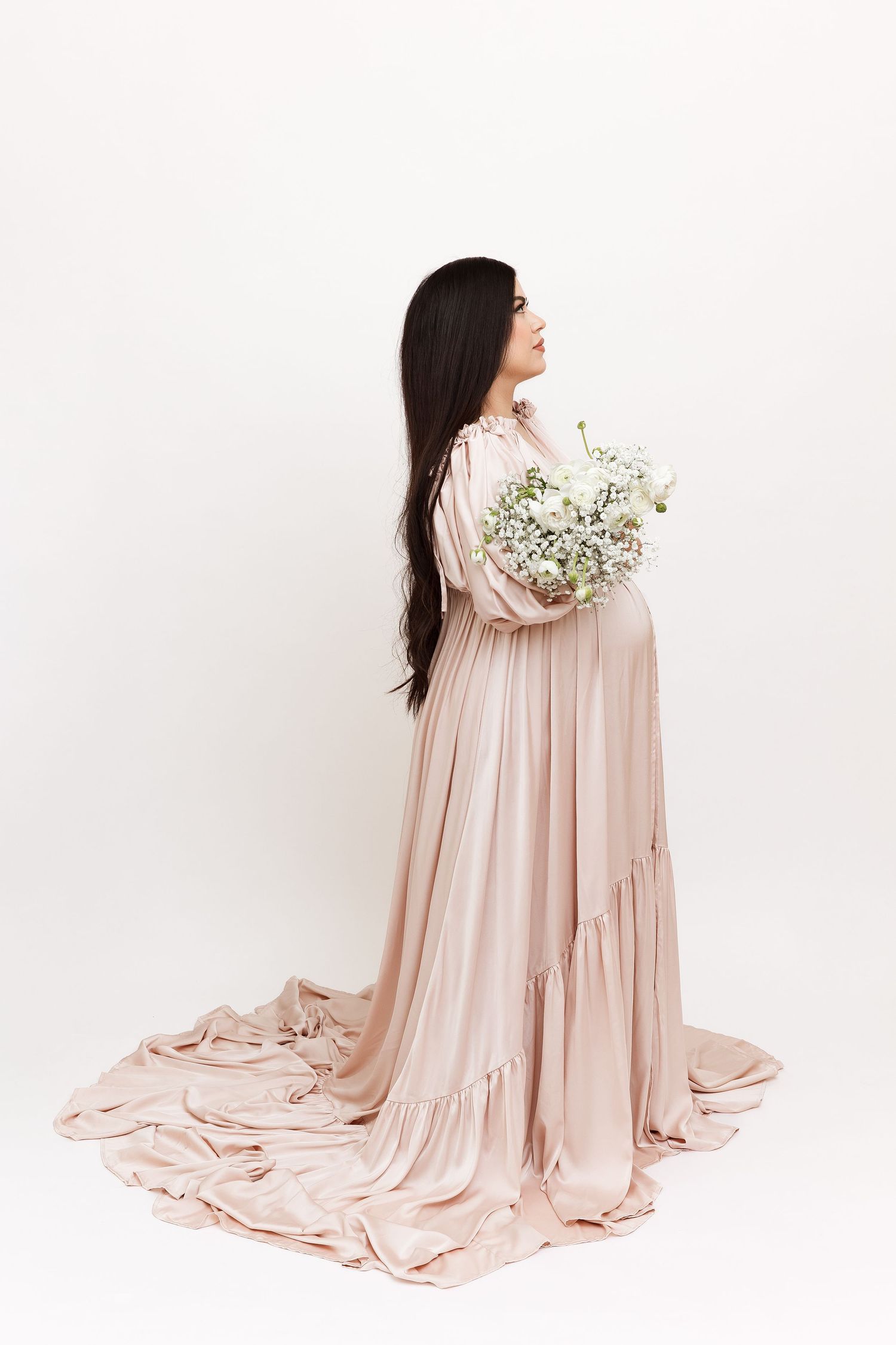 Expectant figure in flowing blush pink gown holding baby's breath flowers against white wall.