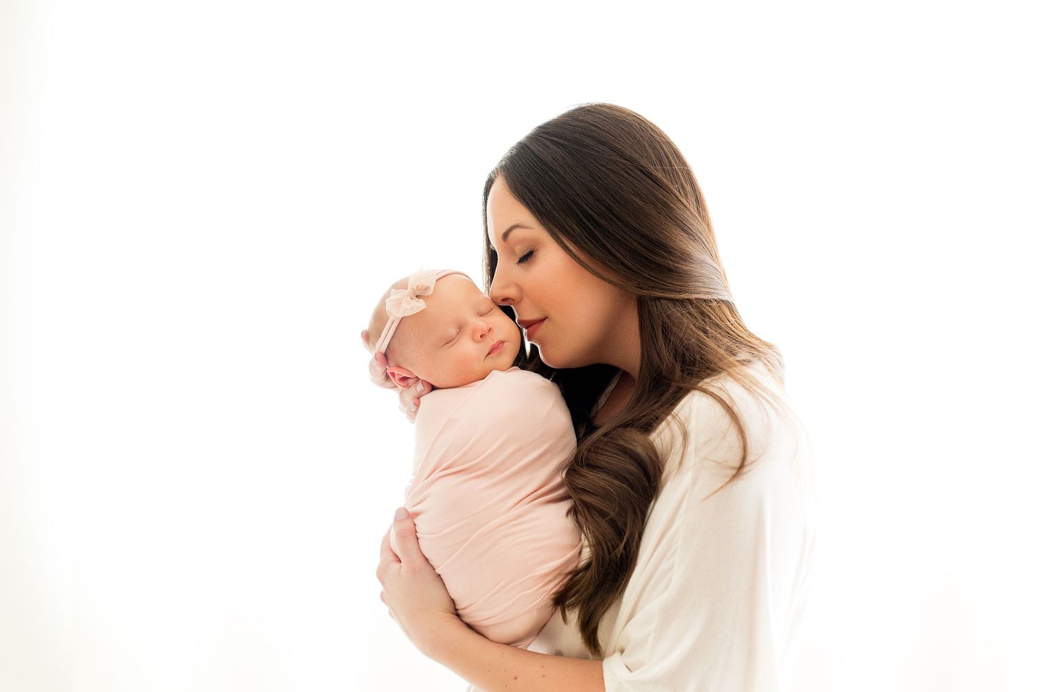 Mother in white top tenderly kissing newborn baby wrapped in pink swaddle.