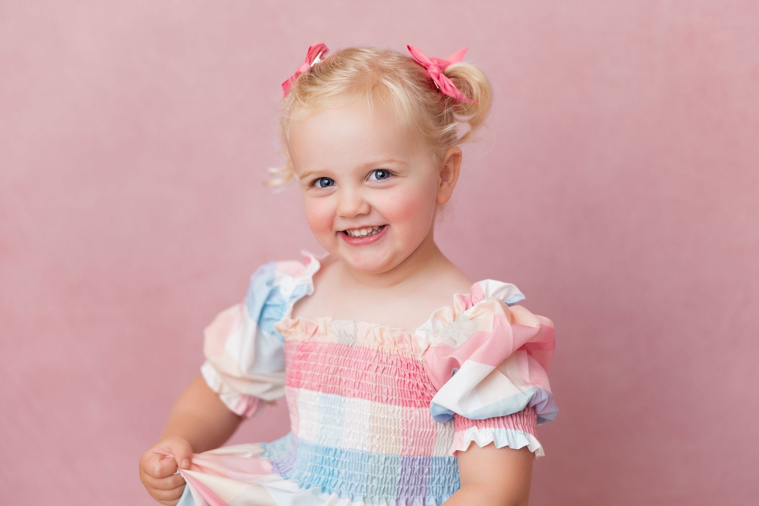 Smiling young child in pastel striped dress with puff sleeves and pink hair bows against pink backdrop.