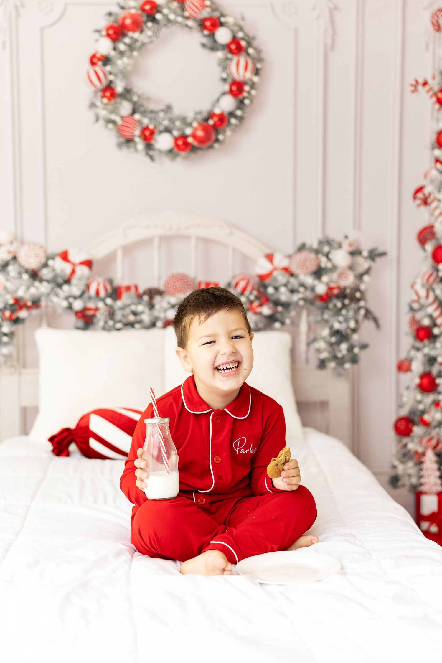 Holiday portrait of child in red pajamas on white bed with festive wreaths.