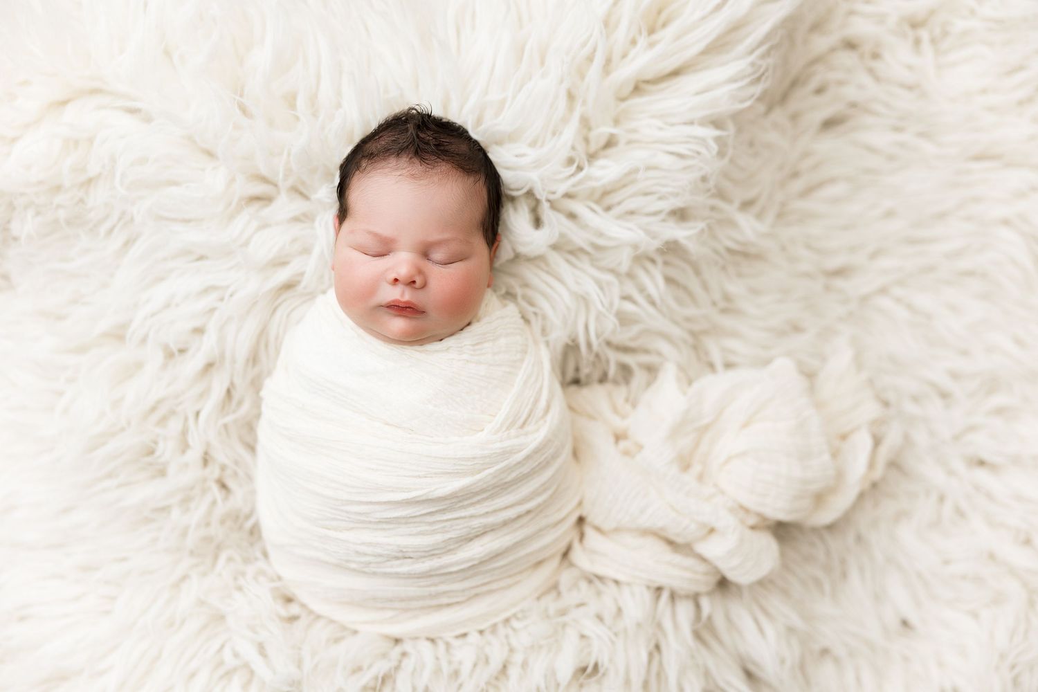 A sleeping newborn baby wrapped in white swaddling cloth lies on a soft white fur blanket.