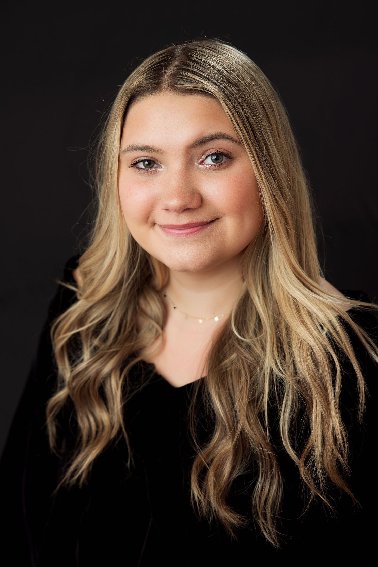 Young woman with long blonde wavy hair smiling, wearing a black velvet top and delicate necklace against dark background.