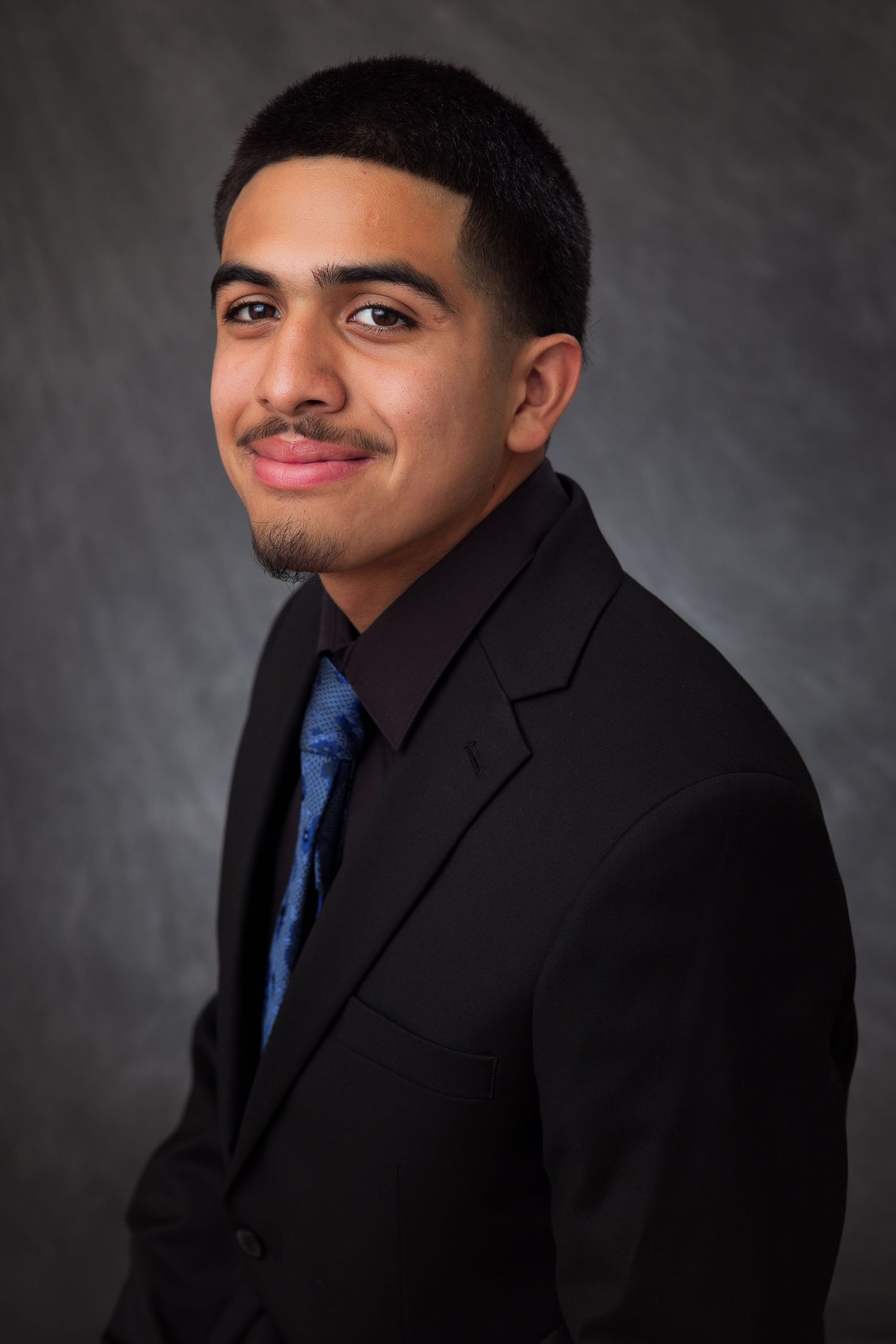 Young professional man in black suit and blue tie smiling in a formal portrait against gray background.