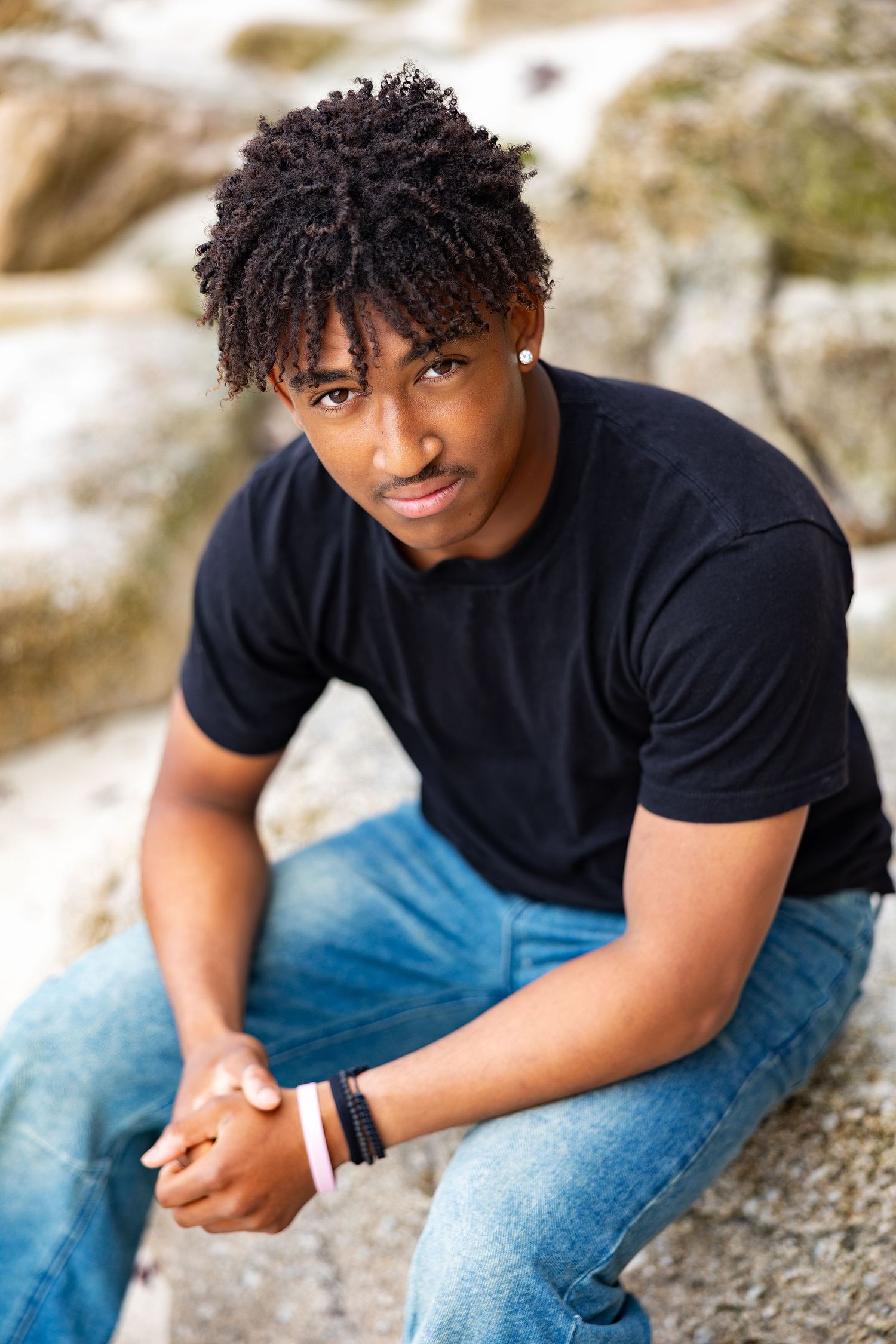 Young Black man in black t-shirt and jeans with curly hair sits on rocks outdoors wearing bracelets.