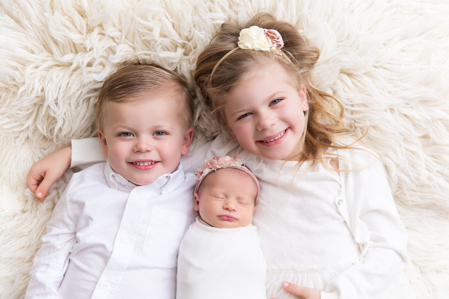 Four children lying together on a soft white fluffy rug, dressed in matching white outfits.