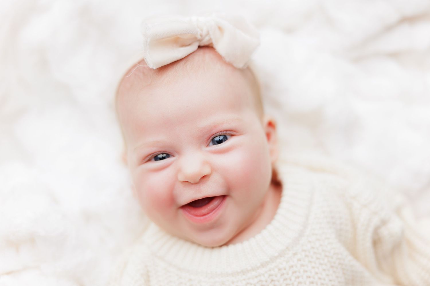 A joyful baby wearing a cozy cream-colored sweater lies on white bedding while smiling brightly.