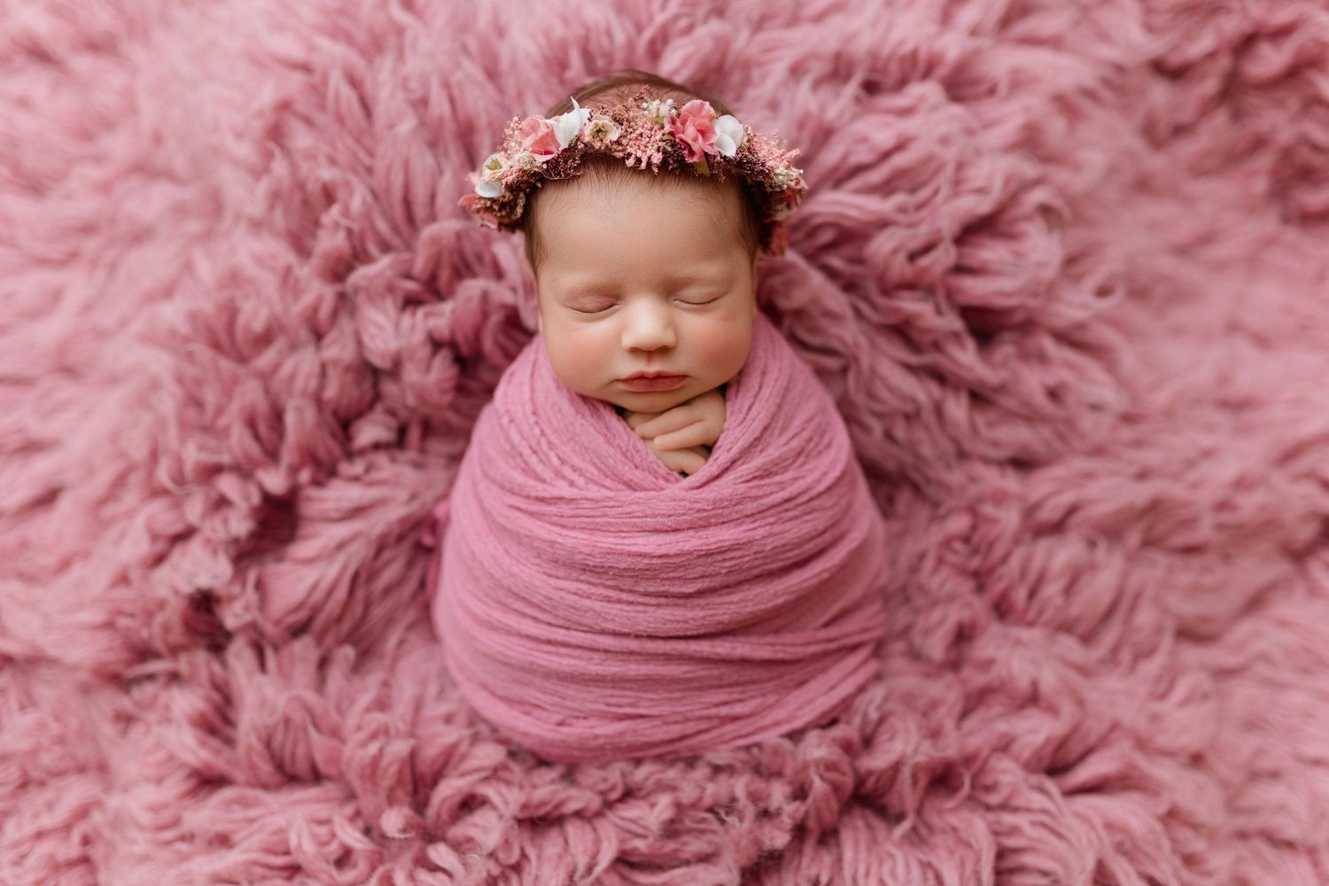 Sleeping newborn wrapped in pink swaddle with floral crown on pink fluffy rug.