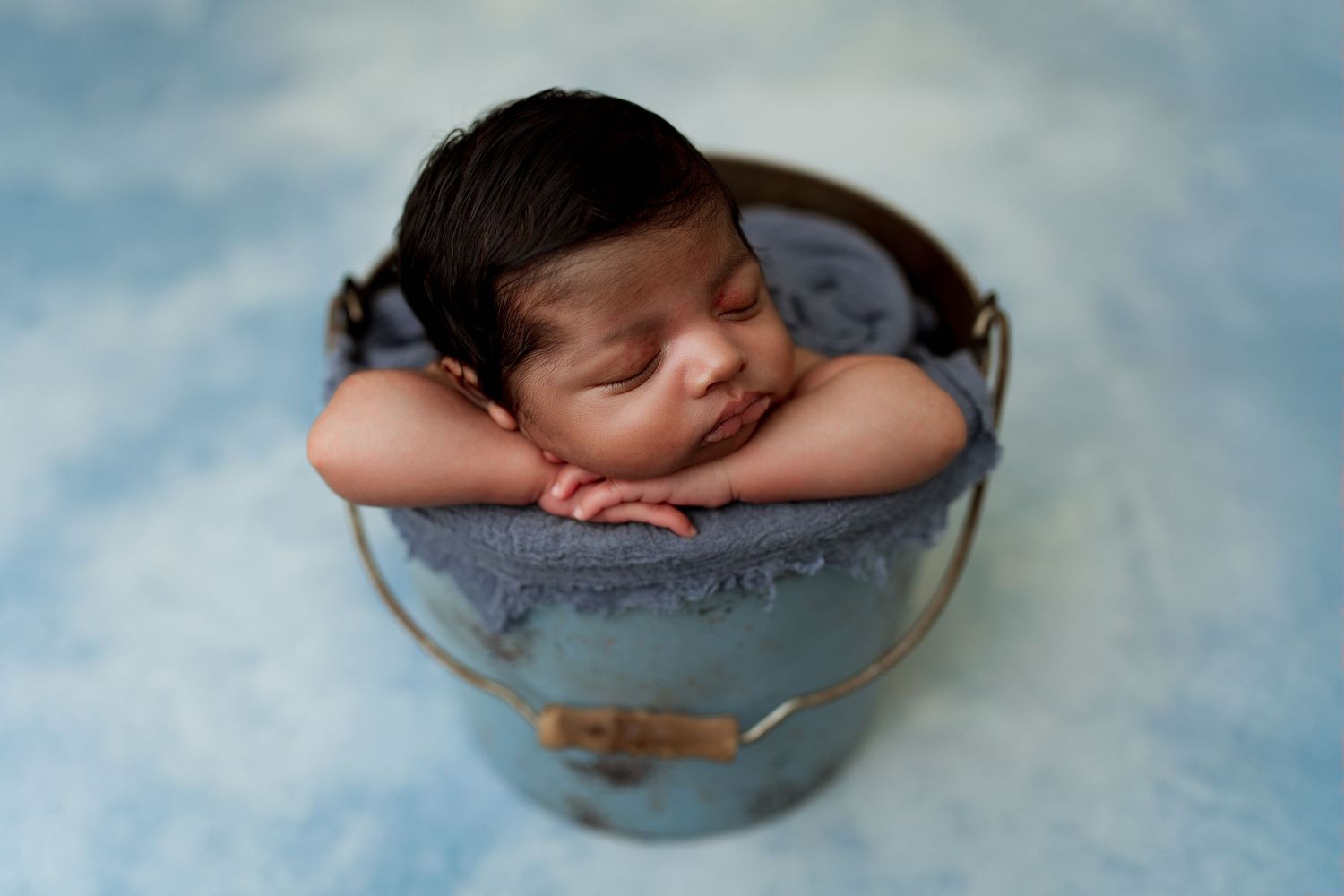 Sleeping newborn posed in vintage blue metal bucket against blue background.