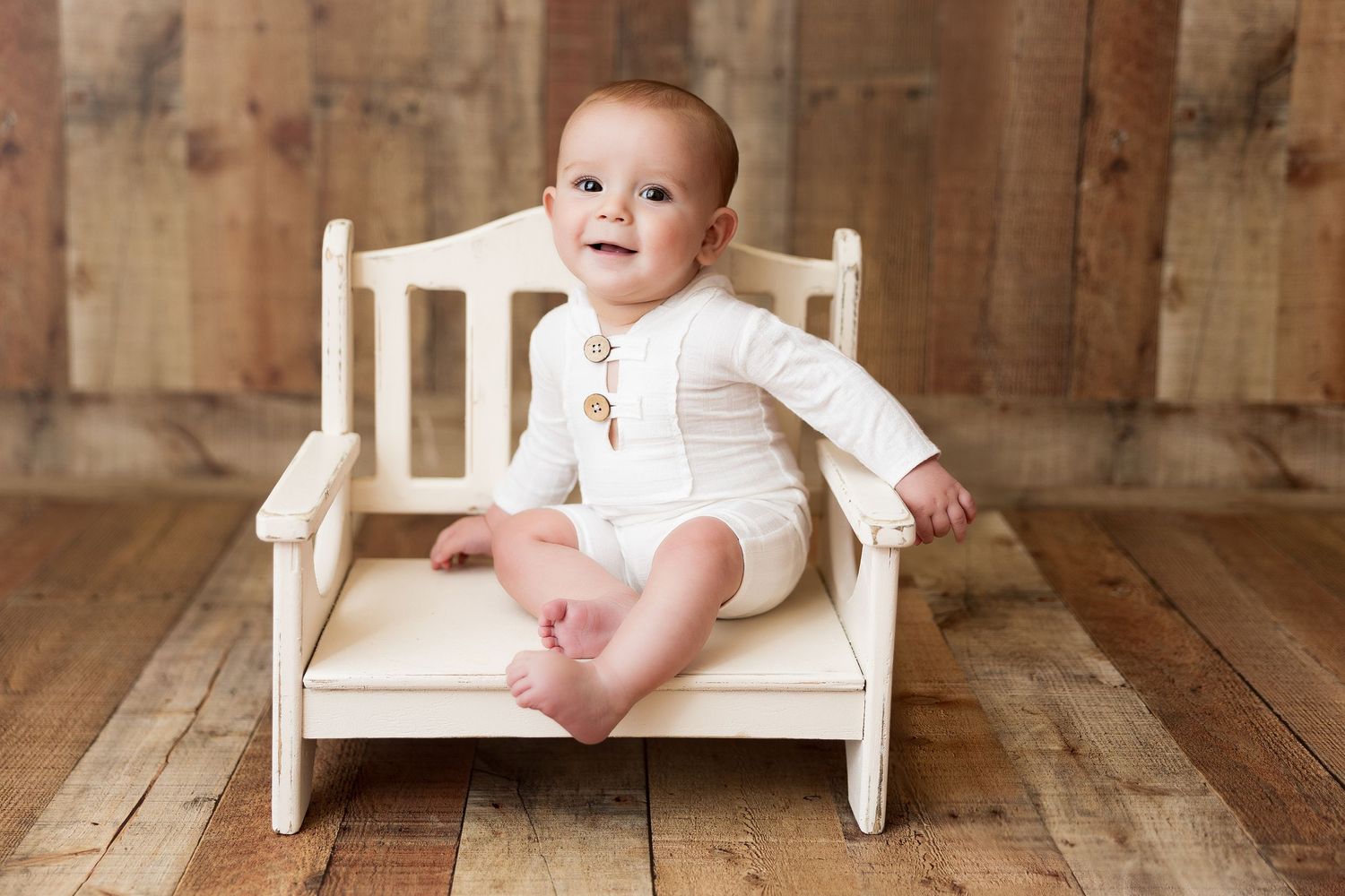Baby sitting on rustic wooden bench against vintage backdrop during milestone photoshoot.