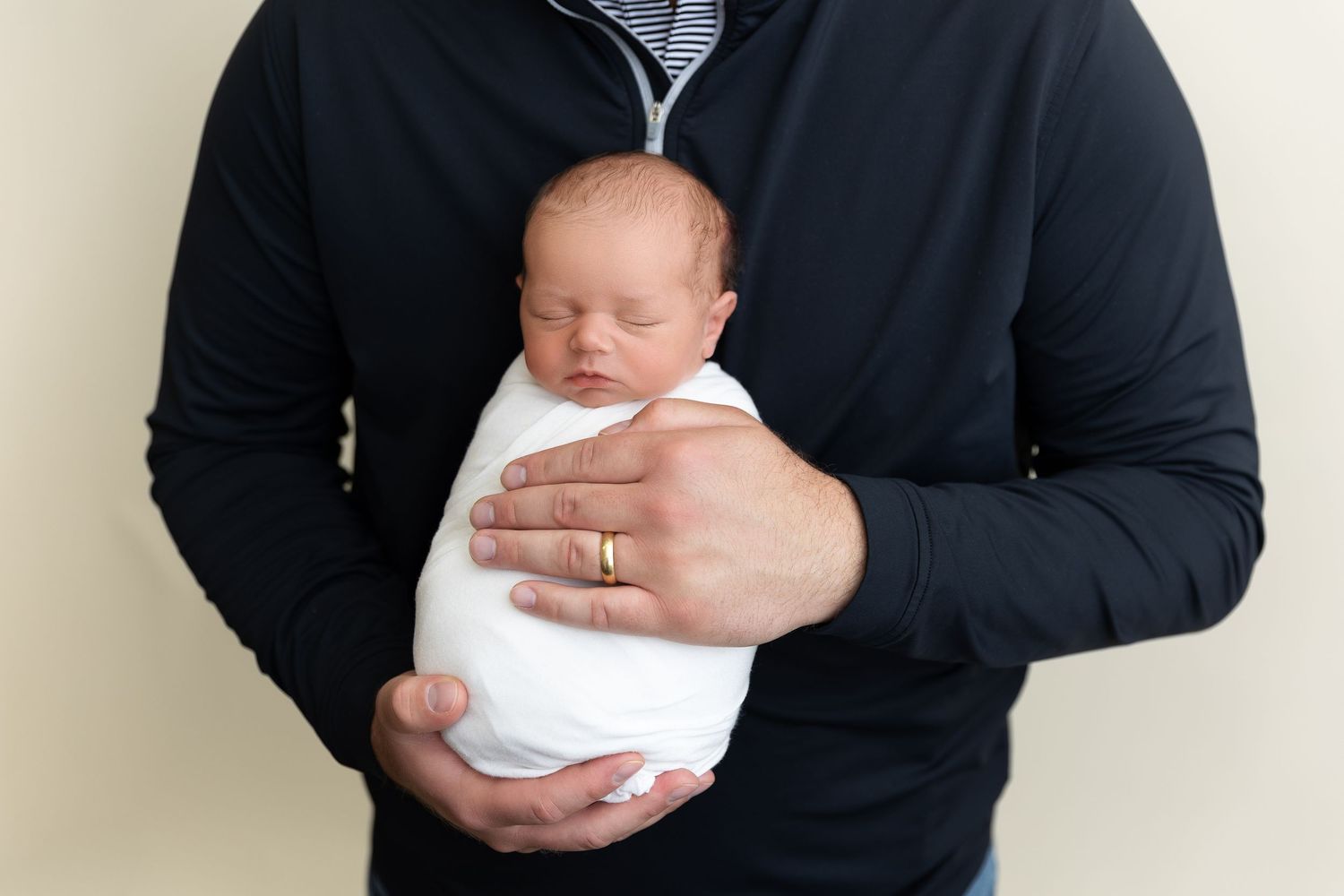 A newborn baby wearing white is cradled securely in someone's arms wearing a dark sweater.