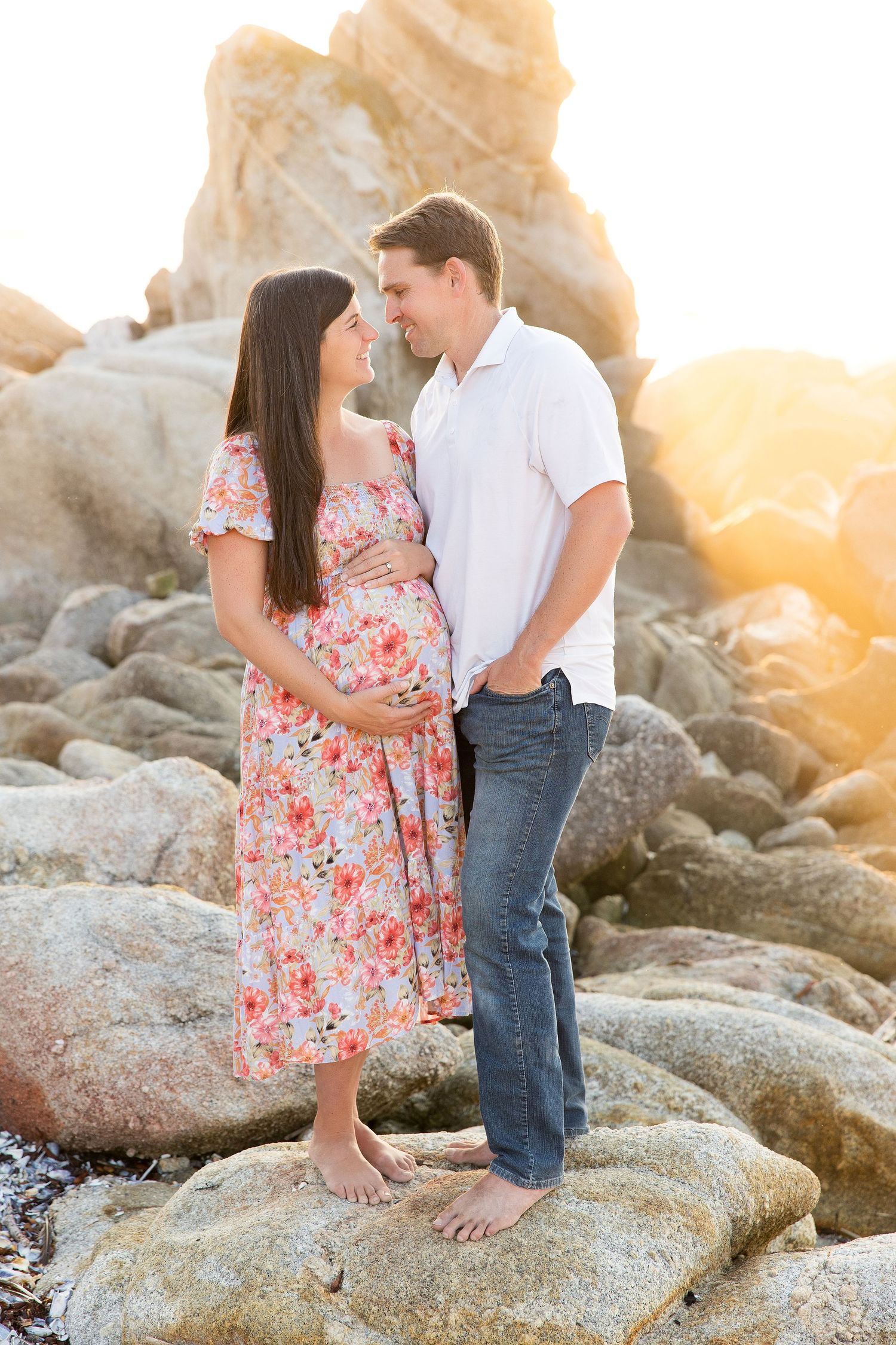 Couple embraces on coastal rocks during sunset maternity photoshoot.