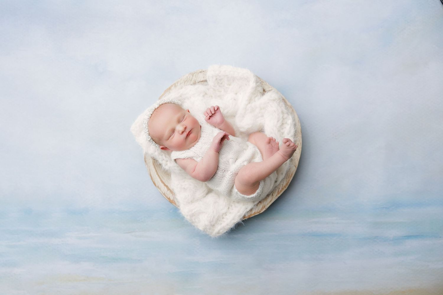 Newborn baby sleeping peacefully in a round wooden bowl with white fur, against a soft blue sky background.