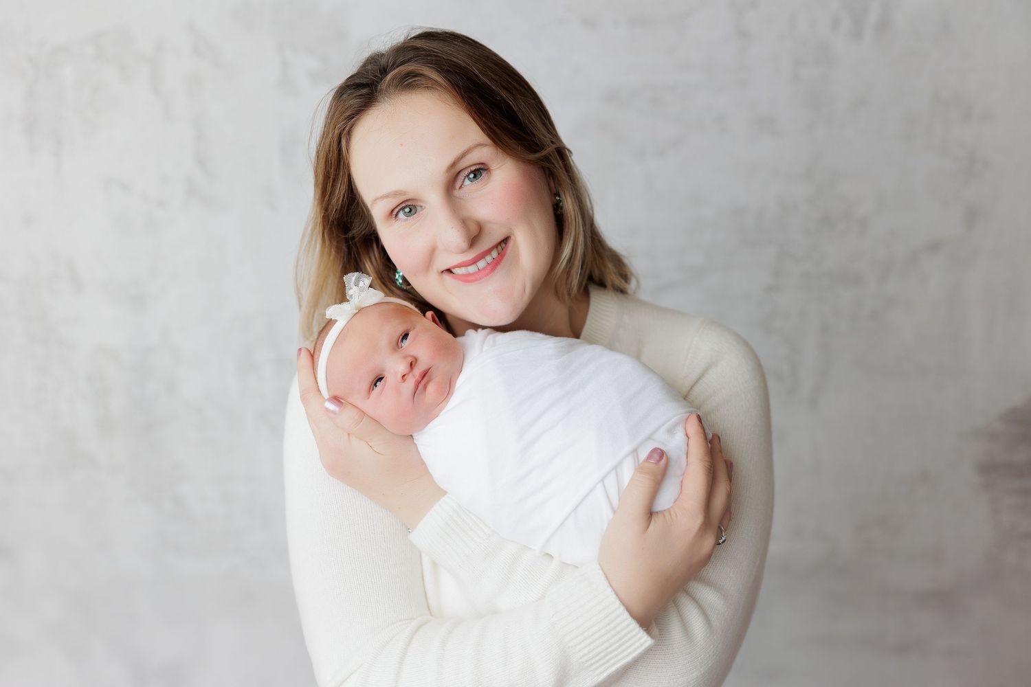 Smiling mother in white sweater holding swaddled newborn baby with white bow against gray background.