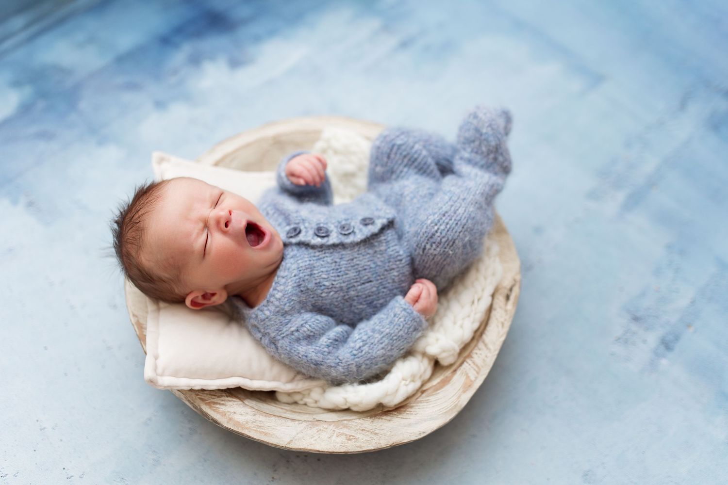 Yawning baby wearing gray sweater lying in woven basket against blue background.