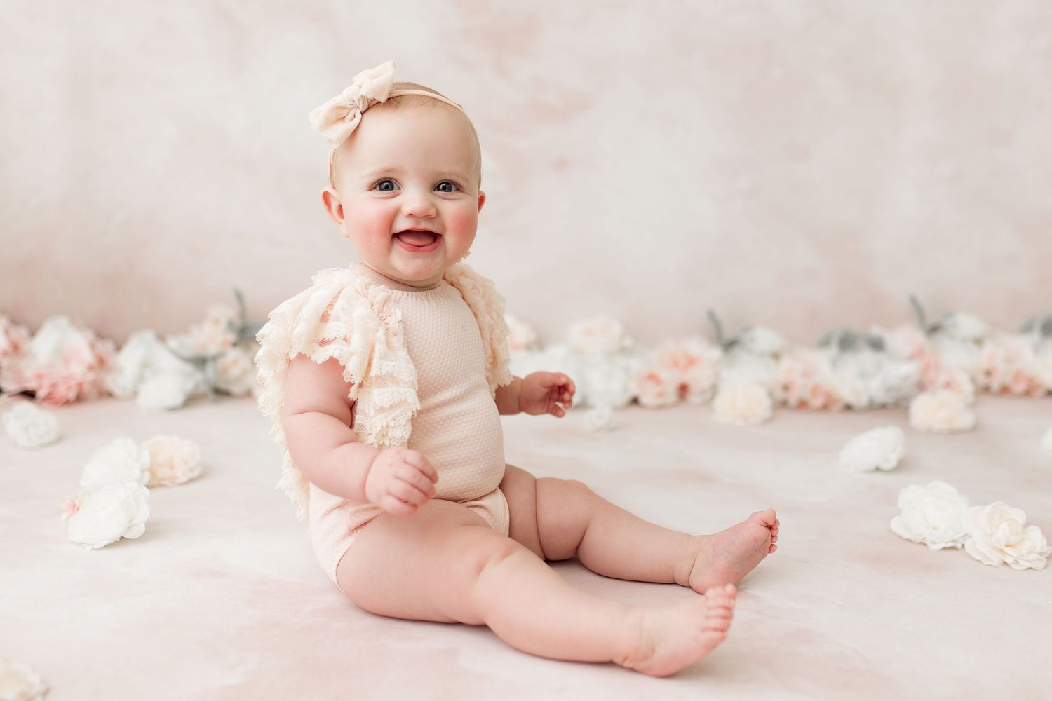 Baby in cream colored outfit sitting on pink background with floral decorations.