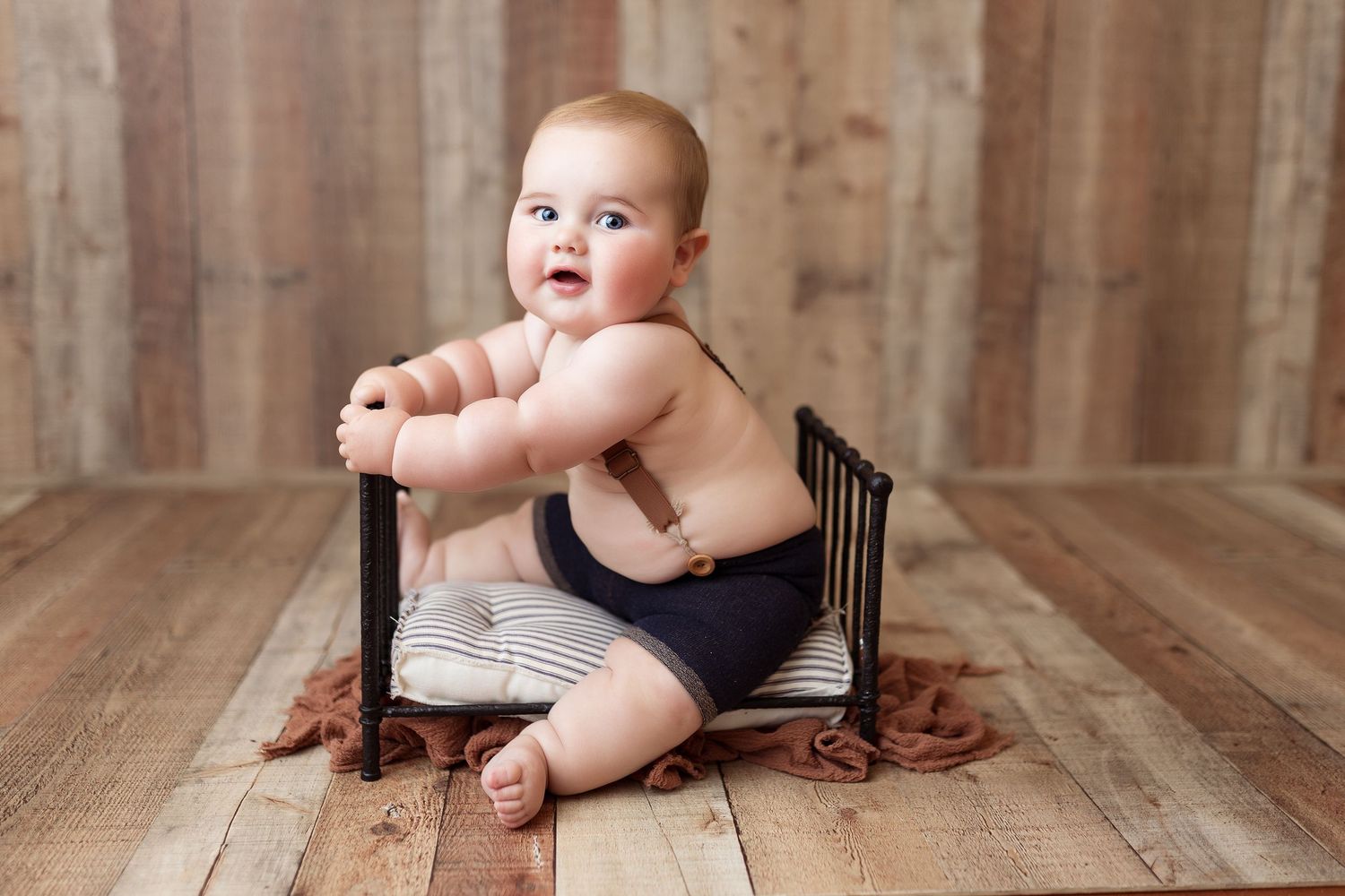 Baby in suspender shorts sits on wooden chair against rustic barnwood backdrop.